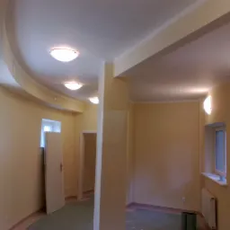 Wnętrze po remoncie: gładkie, beżowe ściany, podwieszany sufit z zaokrąglonymi krawędziami i okrągłymi lampami, widoczne fragmenty glazury i terakoty na podłodze, częściowo zakryte folią ochronną.